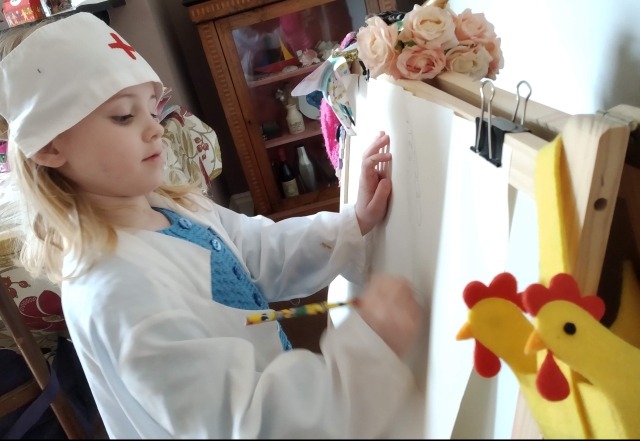 Child in a medical coat and hat writing on an easle